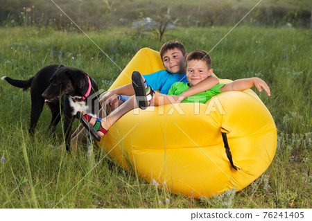 two boys in nature, lie on a yellow lamzak, next to a black dog two boys in nature, lie on a yellow lamzak, next to a black dog 76241405
