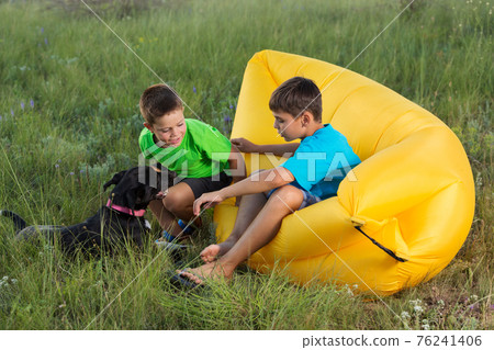two boys outdoors sitting on yellow air couch playing with dog, summer concept two boys outdoors sitting on yellow air couch playing with dog, summer concept 76241406