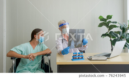 An old woman doctor with a face shield and a protection mask show the Chest X-ray film to the patient sitting in a wheelchair. Describe the symptoms of infectious pneumonia. Covid-19 testing in clinic An old woman doctor with a face shield and a protection mask show the Chest X-ray film to the patient sitting in a wheelchair. Describe the symptoms of infectious pneumonia. Covid-19 testing in clinic 76243945