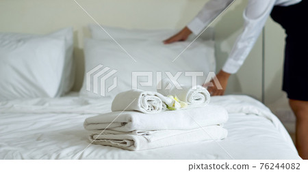 Closeup white towel with white flower on bed. Young hotel maid making the bed in the background. 76244082