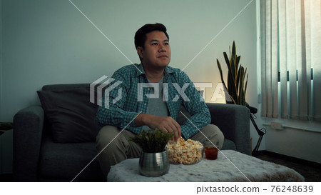 Asian man watching a living room series while eating a popcorn at night. 76248639