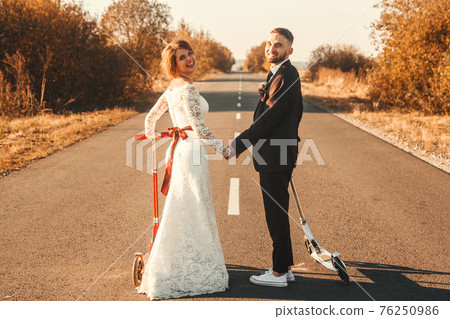 Smiling wedding couple riding a on scooters along the road outside the city at sunset. Smiling wedding couple riding a on scooters along the road outside the city at sunset. 76250986