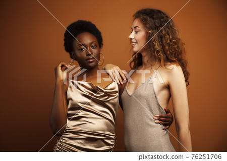 two pretty girls african and caucasian blond posing cheerful together on brown background, ethnicity diverse lifestyle people concept 76251706