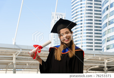 Smiling female in graduation gown holding academic degree and looking at camera with building background. Smiling female in graduation gown holding academic degree and looking at camera with building background. 76253371