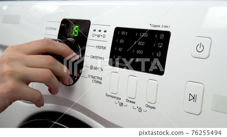 A woman turns the lever choosing the mode on the washing machine and turns it on. Translation: "cotton, synthetics, wool, quick wash, cold, temperature, wring out, options, hold 3 second" 76255494