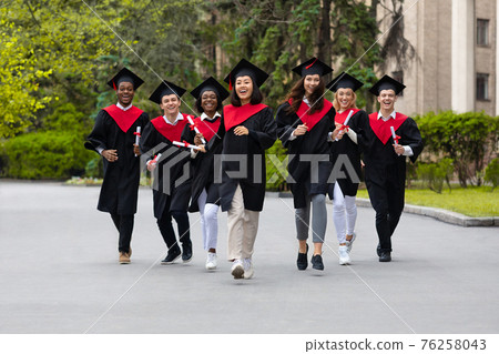 Cheerful multiracial students in graduation costumes walking by university campus 76258043