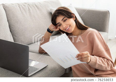 Woman holding paper reading report working on laptop at home Woman holding paper reading report working on laptop at home 76258653