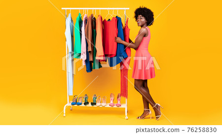 Black Lady Choosing Clothes Standing Near Clothing Rail, Yellow Background Black Lady Choosing Clothes Standing Near Clothing Rail, Yellow Background 76258830