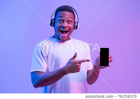 Online Music Platform. Excited African Man In Headphones Pointing At Black Smartphone Online Music Platform. Excited African Man In Headphones Pointing At Black Smartphone 76259379