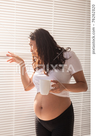 Curious young beautiful pregnant woman drinking tea and looking through the blinds at the window. Concept of joy and good news while waiting for baby 76262820