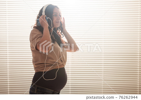 Curly-haired brunette pacified pregnant woman listens to pleasant classical music using smartphone and headphones. Concept of a soothing mood before meeting baby. Copyspace Curly-haired brunette pacified pregnant woman listens to pleasant classical music using smartphone and headphones. Concept of a soothing mood before meeting baby. Copyspace 76262944