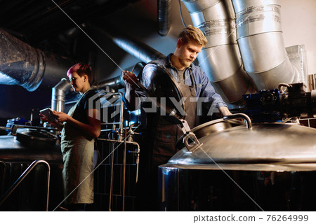 Young woman in workwear using tablet while her male colleague opening lid of huge steel cistern Young woman in workwear using tablet while her male colleague opening lid of huge steel cistern 76264999