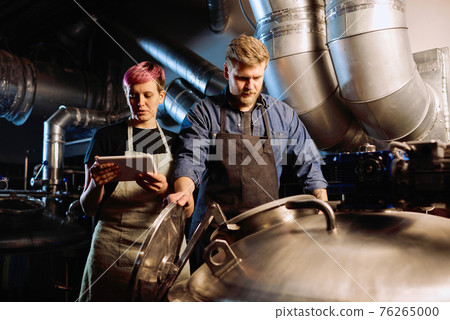 Young woman with digital tablet standing next to male colleague opening lid of huge steel cistern Young woman with digital tablet standing next to male colleague opening lid of huge steel cistern 76265000