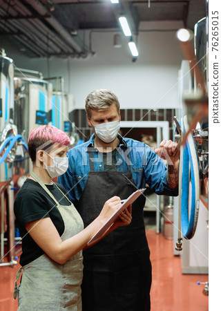 Two young workers of large contemporary beer production factory discussing notes during work 76265015