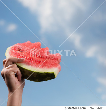 one hand of a small child is raised up, holding a large piece of watermelon, against the blue sky with a cloud, summer concept 76265605