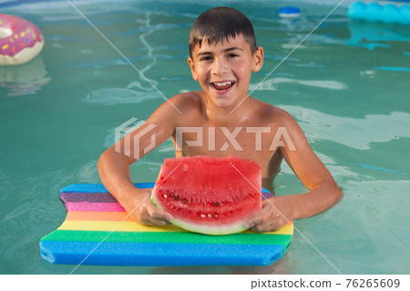 wet boy in the pool with a swimming board, holds in his hands a piece of watermelon, emotions, summer vacation concept wet boy in the pool with a swimming board, holds in his hands a piece of watermelon, emotions, summer vacation concept 76265609