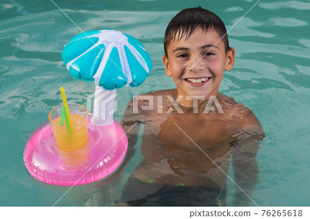 happy little boy smiling, swimming in the pool, next to an inflatable circle with a yellow drink, summer concept 76265618