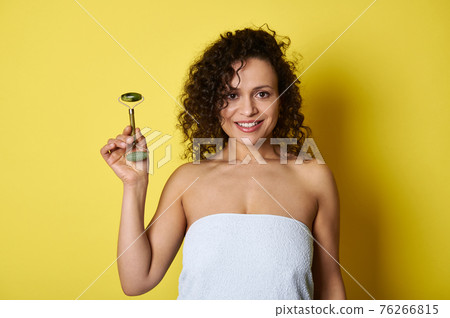 Beauty portrait of young curly haired woman holding jade roller and smiling toothy smile posing over yellow background 76266815