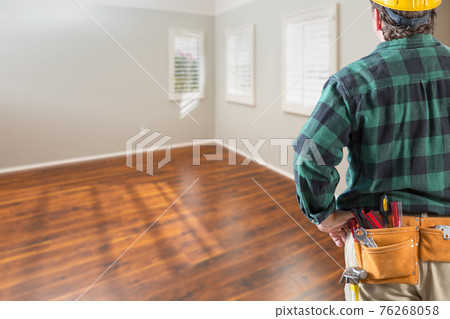 Contractor Wearing Toolbelt and Hard Hat Facing Empty Room with Hard Wood Floors 76268058