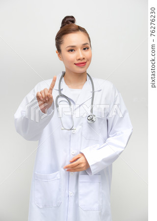 Young beautiful woman doctor in a white coat shows a sign with finger attention on white background. Young beautiful woman doctor in a white coat shows a sign with finger attention on white background. 76276230