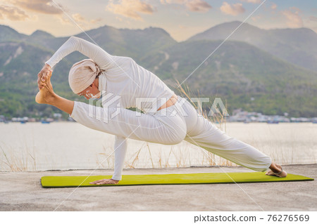 Kundalini yoga woman in white clothes and turban practices yoga kundalini on the background of the sea, mountains and sunset. Fighting face painting of the Indians shows her inner world. Visual 76276569