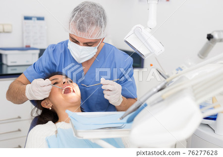 Dentist man examining a latin female patient teeth 76277628