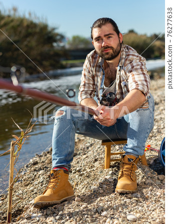 Portrait of cheerful afro fisherman sitting on wooden stool and fishing 76277638