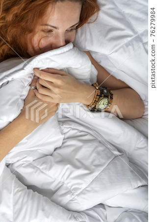 Ginger haired girl shyly peeks out from under the covers. Beautiful young women in her bed 76279984