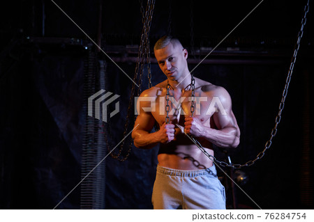 Young shirtless muscular man standing among metal chains, looking at camera, copy space 76284754