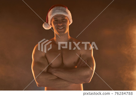 Portrait of muscular man wearing Christmas Santa hat, folded hands on smoky background 76284788
