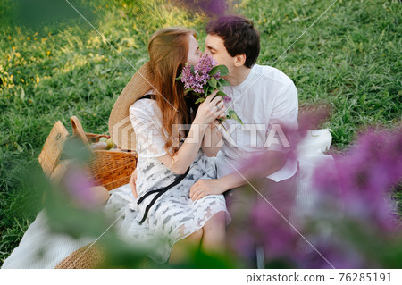 Husband and wife sitting on a light blanket in the park on a picnic kissing closing in with blooming lilacs. 76285191
