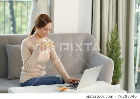 Beautiful cheerful redhead girl using silver laptop while sitting on sofa in living room at home. Beautiful cheerful redhead girl using silver laptop while sitting on sofa in living room at home. 76285340