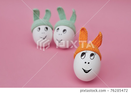 funny egg in orange cap with ears on pink background and two eggs in green caps funny egg in orange cap with ears on pink background and two eggs in green caps 76285472