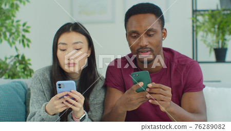 Young mixed ethnicity couple using smartphone while sitting on sofa at home. Couple is spending freetime in social media, using smartphone. Young mixed ethnicity couple using smartphone while sitting on sofa at home. Couple is spending freetime in social media, using smartphone. 76289082
