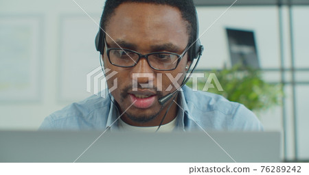 Portrait of African-American man in glasses wears headphones and head microphone uses laptop computer to make online video chat conference online during coronavirus. 76289242
