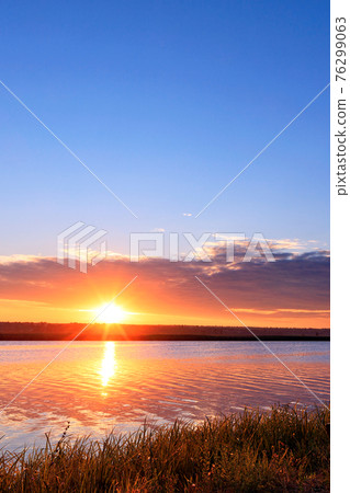 Morning dawn over the river, the bright rising sun rises above the horizon, the sun's rays are reflected in the undulating water surface. Morning dawn over the river, the bright rising sun rises above the horizon, the sun's rays are reflected in the undulating water surface. 76299063