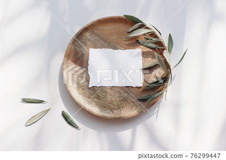 Summer wedding stationery mock-up scene. Blank business card, invitation in sunlight. Wooden plate, tray with olive branches and leaves in sunligh. White table background. Shadows overlay. Flat lay 76299447