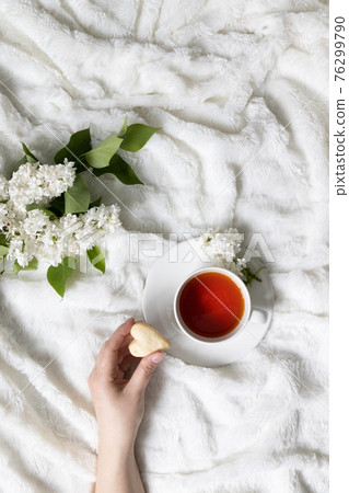Top view of a female hand holding love cookies. Cup of fragrant herbal tea on a white bed frame Top view of a female hand holding love cookies. Cup of fragrant herbal tea on a white bed frame 76299790