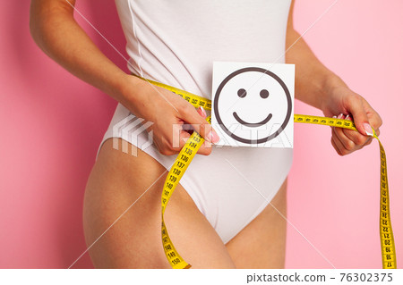 Close up of a woman with a slender figure demonstrates the result holding a card near her stomach with a smiling smile and yellow measure tape Close up of a woman with a slender figure demonstrates the result holding a card near her stomach with a smiling smile and yellow measure tape 76302375
