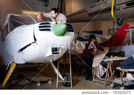 Two young mechanics, man and woman, repairing light sport plane in workshop Two young mechanics, man and woman, repairing light sport plane in workshop 76302675