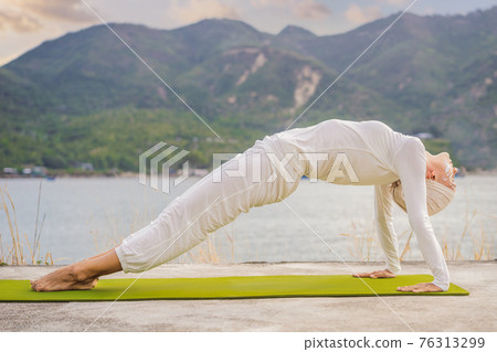 Kundalini yoga woman in white clothes and turban practices yoga kundalini on the background of the sea, mountains and sunset. Fighting face painting of the Indians shows her inner world. Visual 76313299
