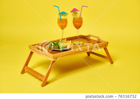 Two glasses with exotic summer cocktails decorated with cocktail umbrella and plate with sliced fruits on bamboo serving tray Two glasses with exotic summer cocktails decorated with cocktail umbrella and plate with sliced fruits on bamboo serving tray 76316732