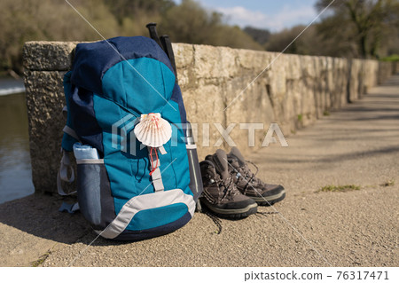 Backpack with seashell symbol of Camino de Santiago, trekking boots and poles leaning on stone wall 76317471
