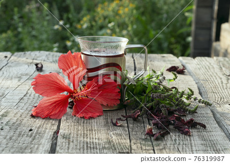 Glass cup of  red herbal Hibiscus tea with ingredients  on wooden table  against a blurred green meadow 76319987