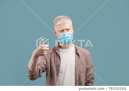 Stay safe. Albino man in medical protective mask showing thumb up on studio background Stay safe. Albino man in medical protective mask showing thumb up on studio background 76320188