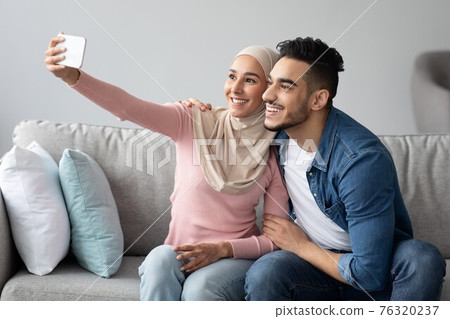 Arab man and woman in hijab smiling at smartphone camera Arab man and woman in hijab smiling at smartphone camera 76320237