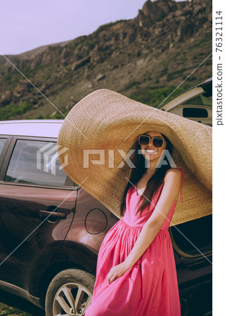 Young beautiful girl in a dress and a wide-brimmed hat near a hatchback car on a background of a mountain landscape. Young beautiful girl in a dress and a wide-brimmed hat near a hatchback car on a background of a mountain landscape. 76321114
