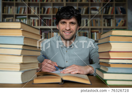 Man student sits at a table in the library, littered with books. Man student sits at a table in the library, littered with books. 76326728
