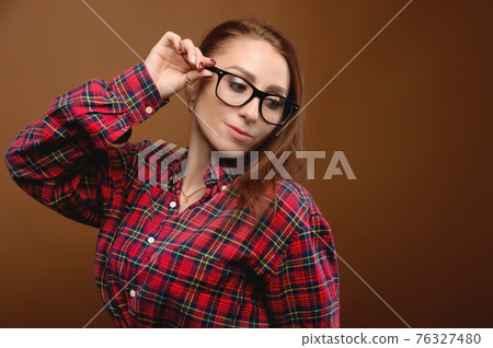 Portrait of a young attractive Caucasian woman in a red shirt and glasses on a brown background. Advertising glasses for improving vision and optics salons 76327480