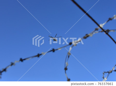 View of the helicopter in the air through the barbed wire against the blue sky View of the helicopter in the air through the barbed wire against the blue sky 76337661
